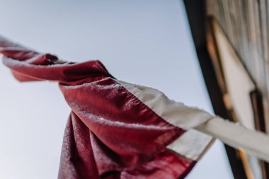 Kırmızı ve beyaz renkte yıpranmış bayrak açık mavi gökyüzüne doğru dalgalanıyor, açık havada kumaşın dokusunu ve hareketini vurguluyor.