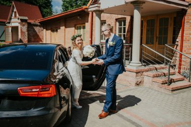 Beyaz elbiseli, çiçekli bir gelin, siyah arabadan iniyor, damat tarafından karşılanıyor, neşe dolu bir düğün havası ve büyüleyici bir mekan sergiliyor.