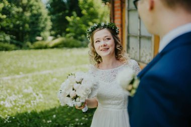 Beyaz elbiseli gelin, çiçekli taçla süslenmiş, neşe içinde damada gülümsüyor, canlı yeşilliklerle dolu güzel bir açık hava ortamında.