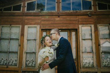 Mutlu çift dışarıda, kırsal ahşap yapının önünde güzel bir anı paylaşıyor, gelin buketi tutuyor ve çiçekli taç takıyor, düğün sevinci ve romantizmi yakalıyor.