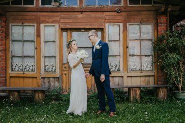 Düğün kıyafetli çift, çimen ve bitkilerle çevrili, klasik ahşap yapının önünde duruyor, doğada bir neşe ve romantizm anı yakalıyorlar.