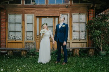 Düğün kıyafetli çift, bereketli çimenler ve çiçeklerle çevrili, eski ahşap yapının önünde sevinçle ayakta duruyor, aşk ve mutluluk anlarını yakalıyorlar.