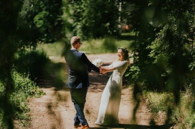 Zarif bir düğün kıyafeti giymiş mutlu çift, resim gibi bir yolda el ele tutuşuyorlar, canlı yeşilliklerle çevrili, bir anlık sevgi ve bağ kuruyorlar.