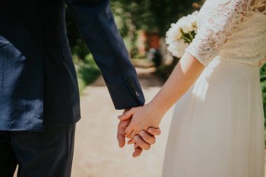 Resmi giyinmiş çift, güzel bir bahçede yürürken el ele tutuşuyorlar, canlı yeşillik ve yumuşak güneş ışığı ile çevrelenmiş, bir anlık sevgi ve bağ kuruyorlar.