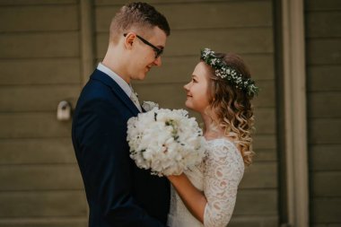 Düğün kıyafetli çift, çiçek buketi ve yeşillikle hassas bir anı paylaşıyor, romantik bir atmosfer yaratıyor, aşkı ve birliği simgeliyor.