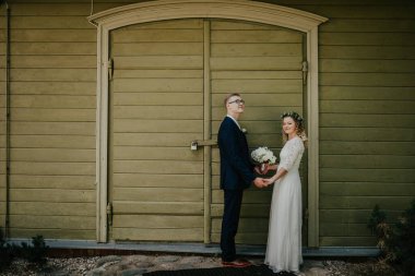 Düğün kıyafetli bir çift, kır yeşil bir kapının önünde duruyor, el ele tutuşuyorlar ve çiçekler, açık havada sevgi ve mutluluk yayıyorlar.