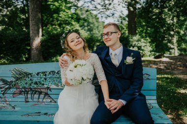 Bir çift, güzel bir düğün atmosferinde, ağaçlar ve doğa ile çevrili, dingin bir parkta, canlı bir bankta, aşklarını kutluyorlar.