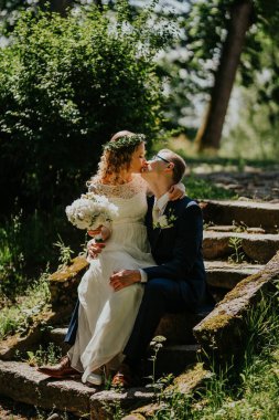 Düğün kıyafetli çift taş basamaklarda oturuyor, canlı yeşilliklerle çevrili, samimi bir anı paylaşıyorlar, doğal bir ortamda sevgi ve neşe sergiliyorlar.