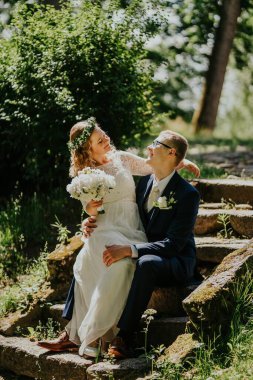 Çift, taştan basamaklarda hassas bir anın tadını çıkarıyor. Etrafı yemyeşil, gelinlik ve çiçeklerle çevrili. Aşk ve neşe çağrıştıran bir taç giymiş gelin.