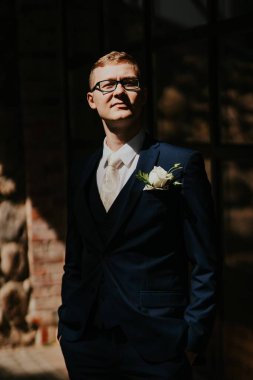 Resmi giyinmiş, gözlüklü, kendine güveni tam genç bir adam yumuşak güneş ışığında duruyor, şık düğün kıyafetini ve beyaz çiçek yaka çiçeğini vurguluyor, sofistike bir atmosfer yaratıyor.