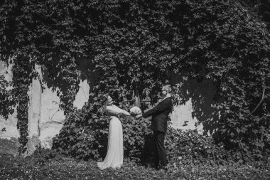 Canlı yeşil bir duvarın önünde el ele tutuşan resmi giyinmiş çift doğa ve aşkla dolu romantik bir atmosfer sergiliyorlar.