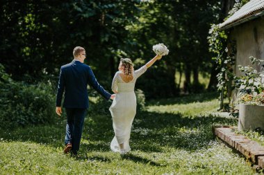 Mutlu çift, canlı bahçede el ele yürür gelin, yeşillik ve doğal güzelliklerle çevrili, neşeli aşk anlarını yakalayan bir buket kaldırır.