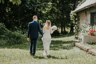 Düğün kıyafetleri içindeki çift canlı yeşilliklerde geziniyor, el ele tutuşuyor ve bir anlık samimiyetin tadını çıkarıyor, arka planda romantik atmosferi yükselten kırsal bir yapı var.