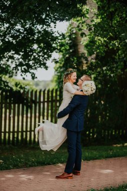 Zarif beyaz gelinlik içinde gelin, damat tarafından donanma elbisesi içinde kaldırılır, yeşil bir zemin ve ahşap bir çite karşı kurulur, sevgi ve kutlama gösterisi sergilenir.