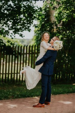 Zarif beyaz elbiseli gelin, damat tarafından canlı yeşillikler ve kırsal ahşap bir çitin arasında kaldırılır, neşe ve kutlama anını yakalar.