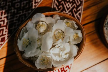 Alyanslar ahşap bir kasenin içindeki yumuşak beyaz çiçeklere yerleştirilmiş, arka planda güzel desenli tekstil, romantik ve zarif bir atmosfer yaratıyor.