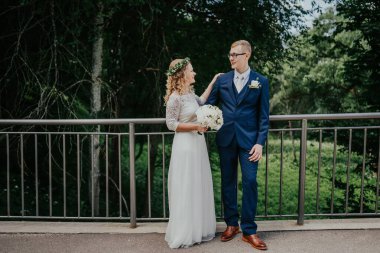 Bir çift köprüde duruyor, gelin buketi tutuyor, damat takım elbise giymiş, canlı yeşilliklerle çevrili, açık havada bir aşk ve mutluluk anı yakalıyorlar.