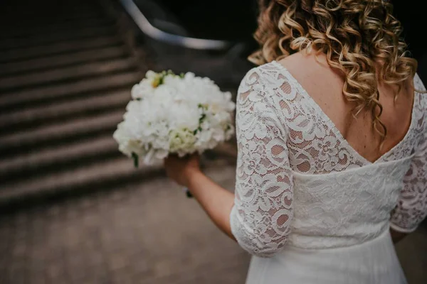 Kıvırcık saçlı gelin, beyaz dantel bir elbise giyiyor, taş basamaklarda duruyor, elinde bir buket beyaz çiçek, güzel bir anda romantizmi ve zarafeti somutlaştırıyor.