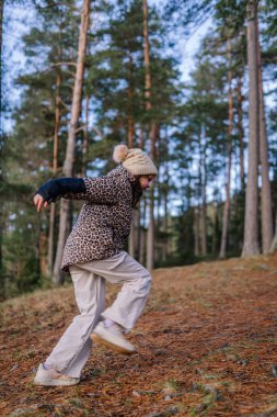 Leopar desenli ceketli ve şapkalı kız uzun ağaçlar ve çam iğnelerinden bir halı ile çevrili bir orman patikasında neşe dolu bir anı yakalıyor.