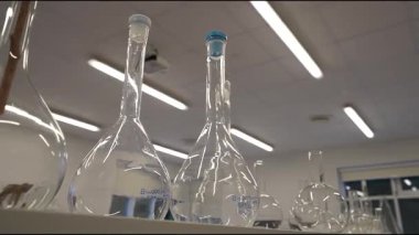 Laboratuvar sahnesinde cam mataralar ve beher kapları bir rafta, şekillerini ve yansımalarını vurguluyor, kamera ise daha yakından görmek için yavaşça yakınlaşıyor.