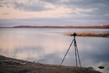 Gün batımında sakin gölün yanında duran tripod üzerine monte edilmiş kamera, yumuşak renkleri ve doğal manzarayı yansıtıyor, açık hava fotoğrafçılığını ve ekipmanları ön plana çıkarıyor.