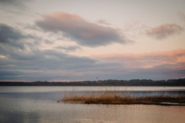 Gün batımında sakin göl manzarası, sakin sular ve pastel bulutlar, kıyı şeridi boyunca uzun otlar, barışçıl bir açık hava atmosferi çağrıştırıyor.