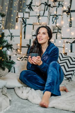 Mavi takım elbiseli kadın, lüks halıda sıcak içecekten hoşlanır, bayram hediyeleri ve parıldayan ışıklarla çevrili, samimi ve neşeli bir ortam uyandırır.