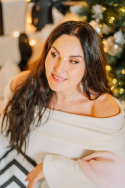 Uzun siyah saçlı bir kadın, rahat bir süveter giymiş, Noel ağacının yanında vakit geçiriyor, sıcak ışıkları parlıyor, neşeli ve davetkar bir tatil ortamı yaratıyor.