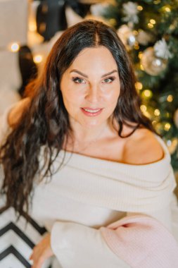 Uzun siyah saçlı, yumuşak bir kazak giymiş bir kadın, güzel dekore edilmiş bir Noel ağacının yanında poz veriyor, neşeli bir ortamda sıcaklık ve neşe saçıyor.