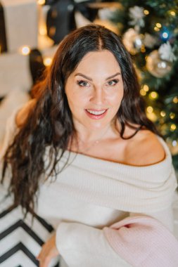 Uzun siyah saçlı, rahat bir süveter giymiş güzel dekore edilmiş bir Noel ağacının önünde gülümseyen, sıcak ve davetkar bir tatil ortamı yaratan bir kadın.