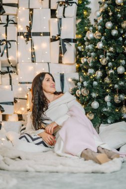 Koyu renk saçlı, kalın elbiseli, şık halıda güzel süslenmiş Noel ağacının yanında rahatlayan ve neşeli tatil havalarını kucaklayan bir kadın figürü.