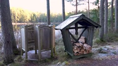 Bir ahşap depolama kulübesi ve açık hava tuvaleti de dahil olmak üzere kırsal ahşap yapılar, doğal güzelliği yansıtan pürüzsüz bir kamera tavasıyla huzurlu bir orman sahnesinde vurgulanır.