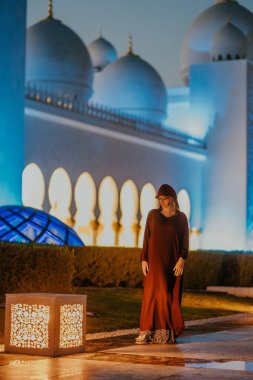 Burgonya elbiseli kadın figürü süslemeli fenerin yanında duruyor, arka planda güzel bir mimari ve yumuşak ışıklandırma, sakin bir akşam sahnesini çağrıştırıyor