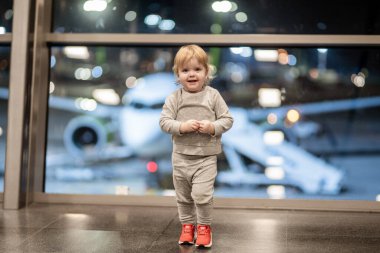 Gri elbiseli ve kırmızı ayakkabılı çocuk havaalanı terminalinde duruyor. Arka planda hafif ışıklandırma ve bulanık uçakla çevrili. Seyahat heyecanı ve masumiyeti yakalıyor.