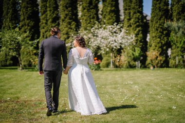 Ogre, Letonya - 11 Mayıs 2024 - Gelin ve damat, güneşli bir günde yeşil bir bahçede el ele yürürler, romantik ve huzurlu bir düğün anında arkadan yakalanırlar..