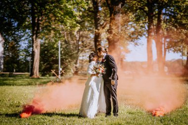 Düğün kıyafetleri içinde romantik bir çift yeşil bir parkta öpüşüyor, etraflarını renkli dumanlar sarıyor, özel günleri için büyülü bir atmosfer yaratıyorlar.