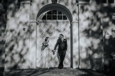Resmi nikah kılığındaki çift, büyük ahşap bir kapının yanında el ele tutuşuyorlar, yumuşak gölgeler romantik bir atmosfer yaratıyor, aşkı ve birliği kutluyorlar.