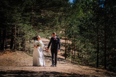 Düğün kıyafetleri içinde çiftler, ağaçların arasından süzülen güneş ışığıyla huzurlu bir orman yolunda yürürler. Özel günleri için romantik ve neşeli bir atmosfer yaratırlar.