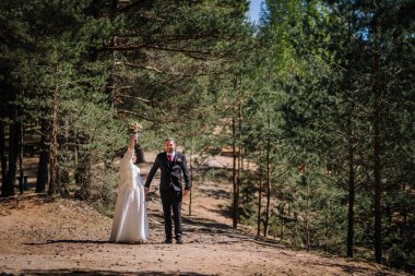 Düğün kıyafetleri içindeki çift, sakin ormanda, damat gelinin elini tutuyor, buketi neşeyle kaldırıyor, yemyeşil ve doğal güzelliklerle çevrili.