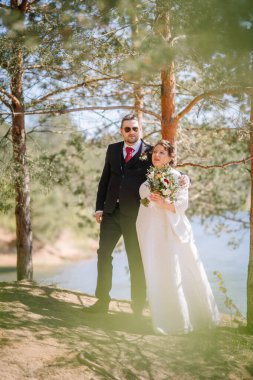 Düğün kıyafetli çift, yemyeşil bir ormanda poz veriyor ve arka planda romantizmi ve birliği simgeleyen sakin bir göl var.