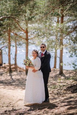 Düğün kıyafetli bir çift, yeşil ve güneş ışığıyla çevrili, sakin bir ormanda duruyor, ellerinde canlı bir buket, aşkı ve birliği simgeliyor.