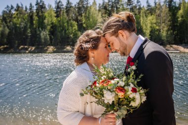 Romantik çift sakin bir gölün kenarında samimi bir anı paylaşıyor. Arka planda canlı bir yeşillik, düğün kutlamaları sırasında sevgi ve neşe sergiliyor.