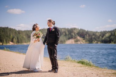 Düğün kıyafetli çift sakin bir gölün yanında duruyor, arka planda canlı yeşillikler, güzel bir günde aşk ve mutluluk anlarını yakalıyorlar.