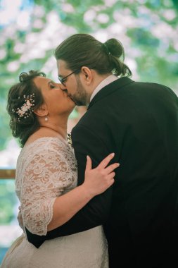 Asyalı çift düğün töreninde hassas bir öpücükle kucaklaşır. Etrafı çiçeklerle çevrilidir. Romantik ve samimi bir ortam yaratır.