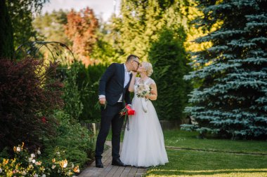 Zarif düğün kıyafetleri içindeki romantik çift güzel bir bahçede bir öpücük paylaşıyor, etrafları renkli çiçekler ve yeşilliklerle çevrili, aşk ve neşe dolu bir anı yakalıyorlar.