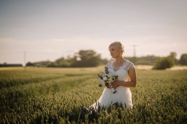 Beyaz elbiseli zarif gelin canlı yeşil bir alanda duruyor, elinde güzel bir buket, sakin bir açık havada romantizm ve neşe var.