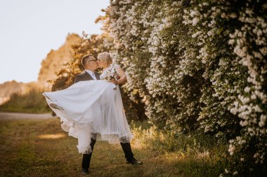 Aşkı kutlayan çift, zarif bir elbise içinde gelin ve damat, yemyeşil ve canlı çiçekler arasında kucaklaşıp neşeli bir atmosfer yaratıyor.