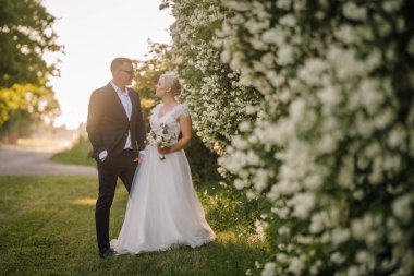 Düğün kıyafetli çift, canlı çiçeklerle çevrili güzel bir bahçede duruyor. Düğün törenlerinde yumuşak akşam ışığında romantik bir anın tadını çıkarıyorlar.