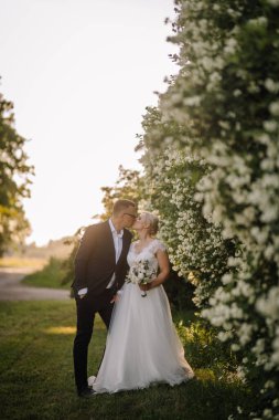 Bereketli yeşilliklerin ve çiçek açan çiçeklerin arasında öpüşen çift, açık havada sakin bir düğün atmosferinde aşkı ve bağlılığı simgeliyor.
