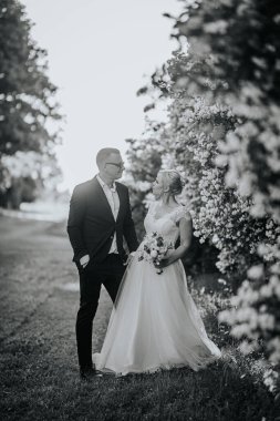 Resmi giyinmiş gelin ve damat canlı çiçeklerle çevrili güzel bir bahçede duruyor ve özel günleri için romantik bir ortam yaratıyorlar.